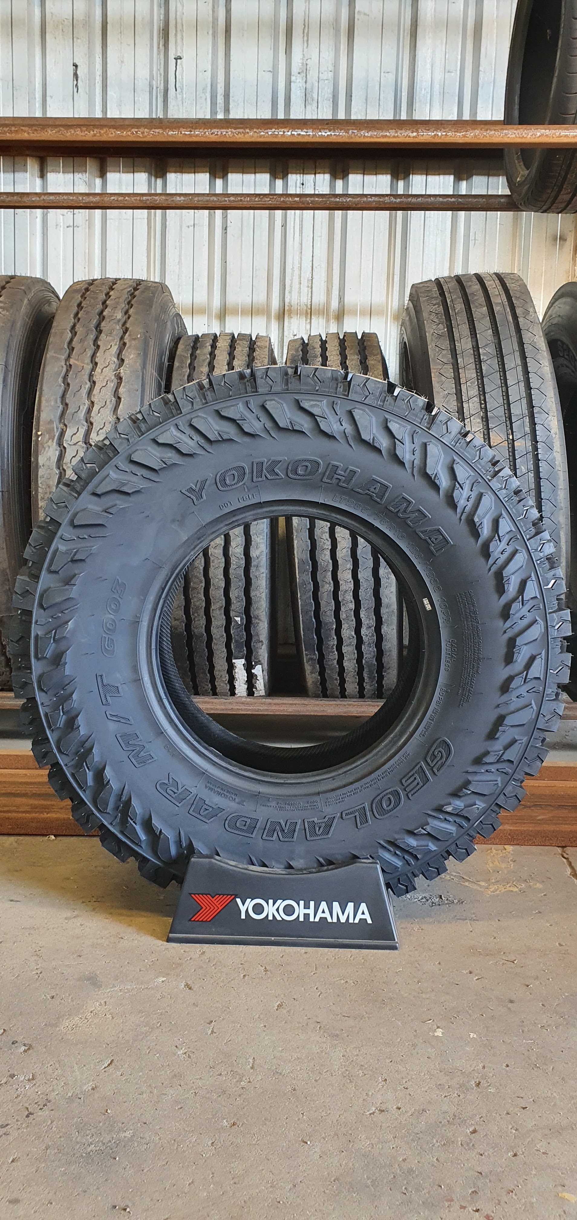 A Yokohama Geolandar M/T G003 tyre on a branded display stand in front of a rack of truck tyres