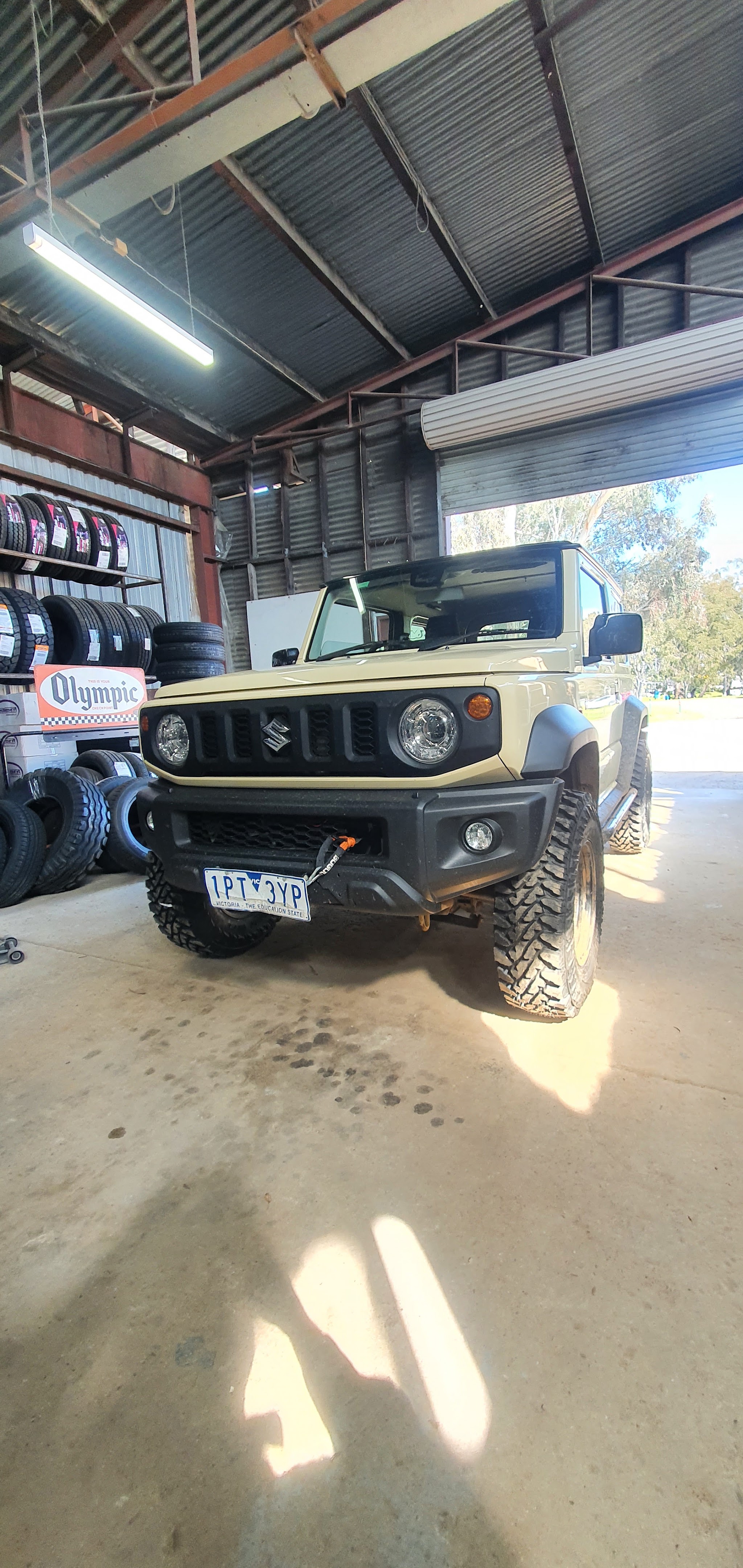 A Suzuki Jimny on new mud terrain tyres inside the workshop, with the roller door open and racks of tyres behind